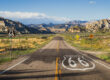 A long stretch of Route 66 runs through the mountains with a large historic highway marker.
