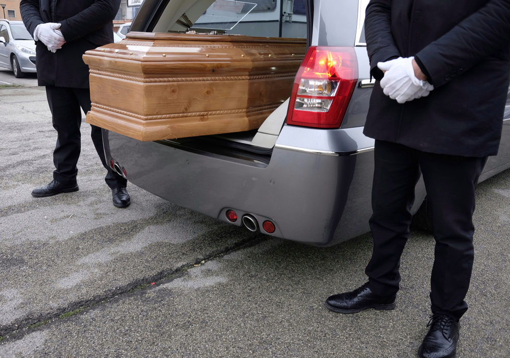 Pallbearers loading a wooden casket into a silver funeral car during a service